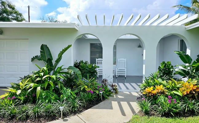 Modern white house with arched openings, pergola, and tropical landscaping with flowering plants and banana trees
